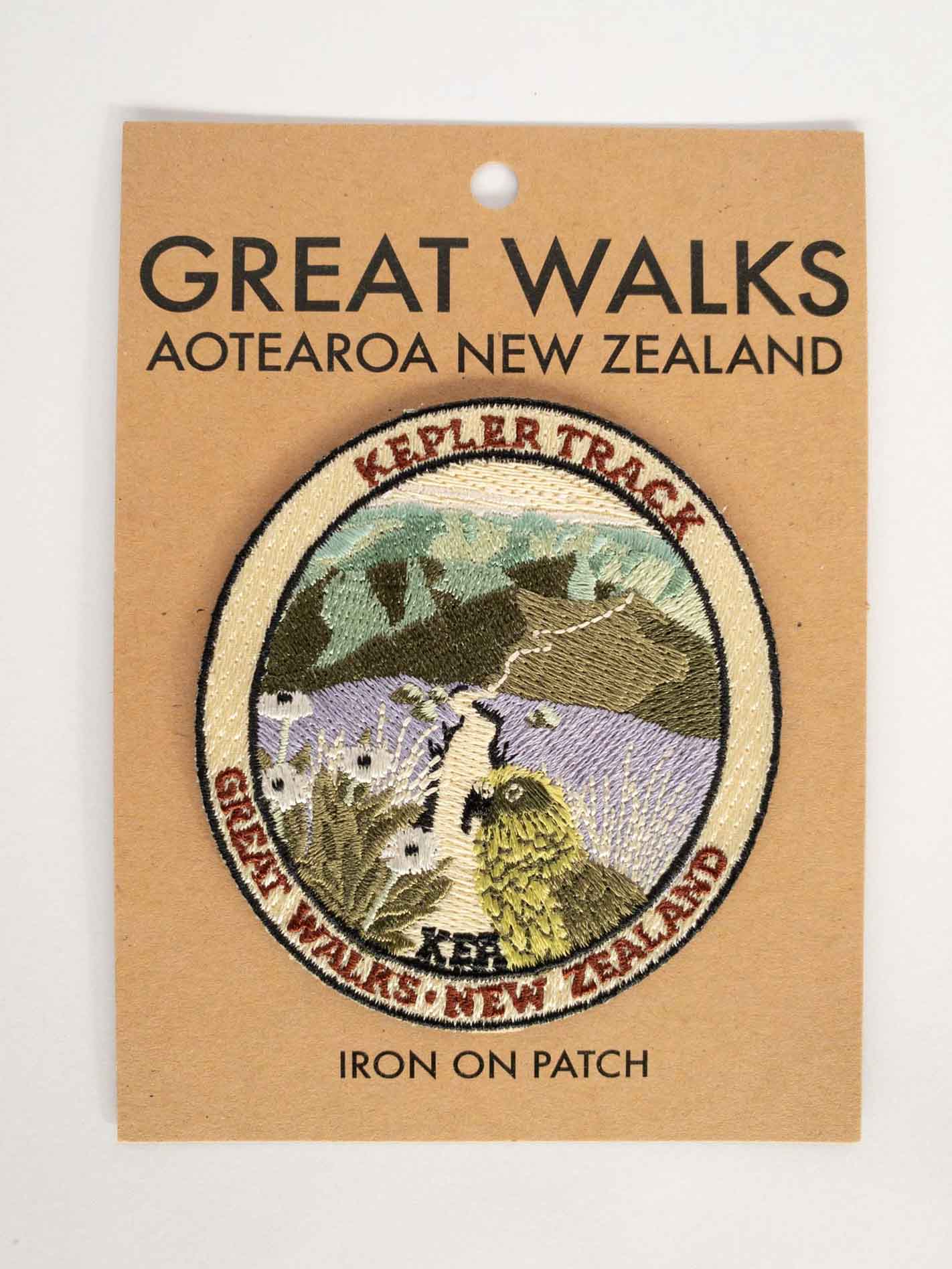 Oval, embroidered Kepler Track patch, with a kea, mountain daisy and alpine ridges, on brown kraft backing card.