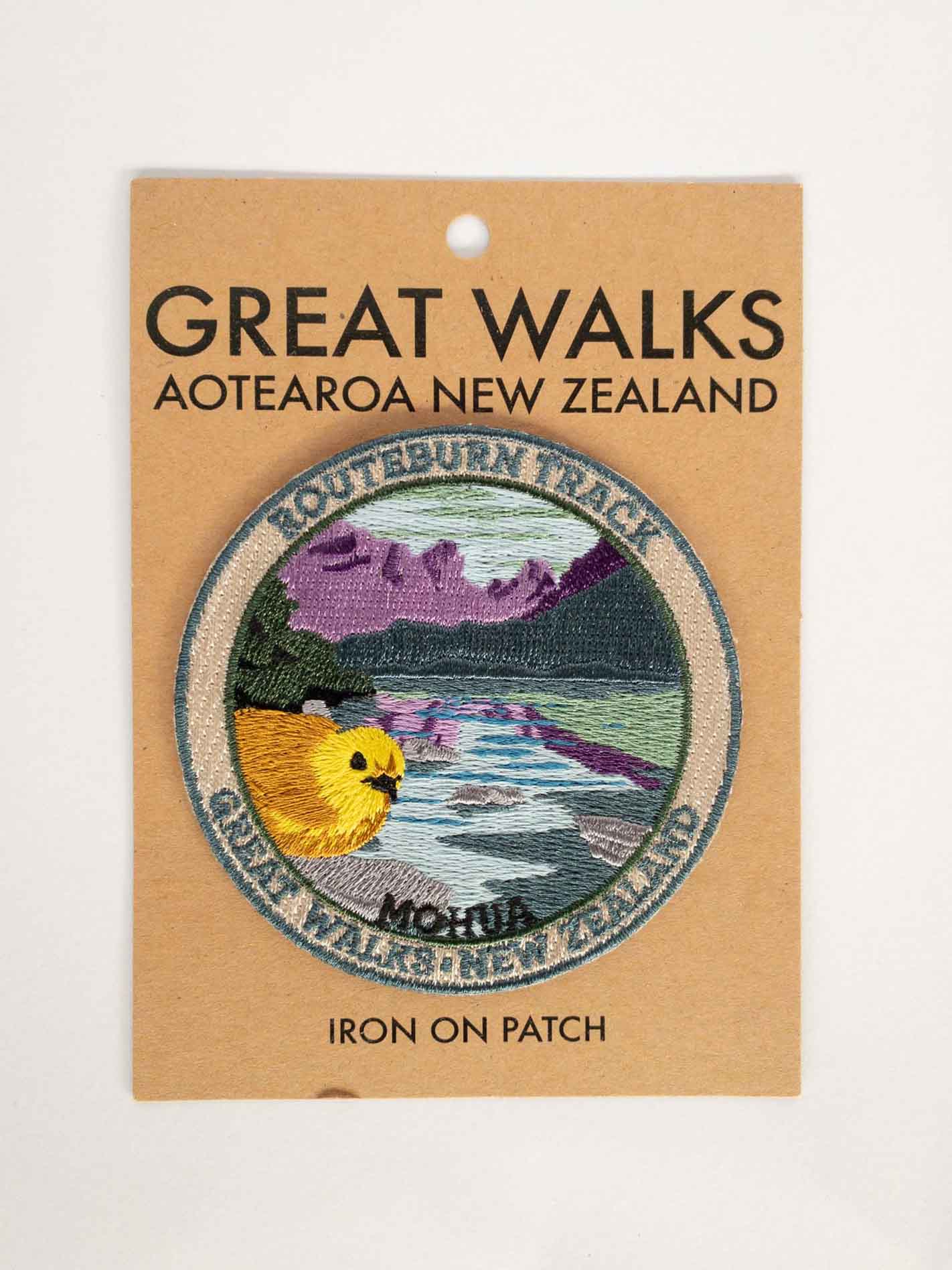 Round, embroidered Routeburn Track patch, with a mohua/yellowhead bird, purple mountains and blue lake, on brown kraft backing card.