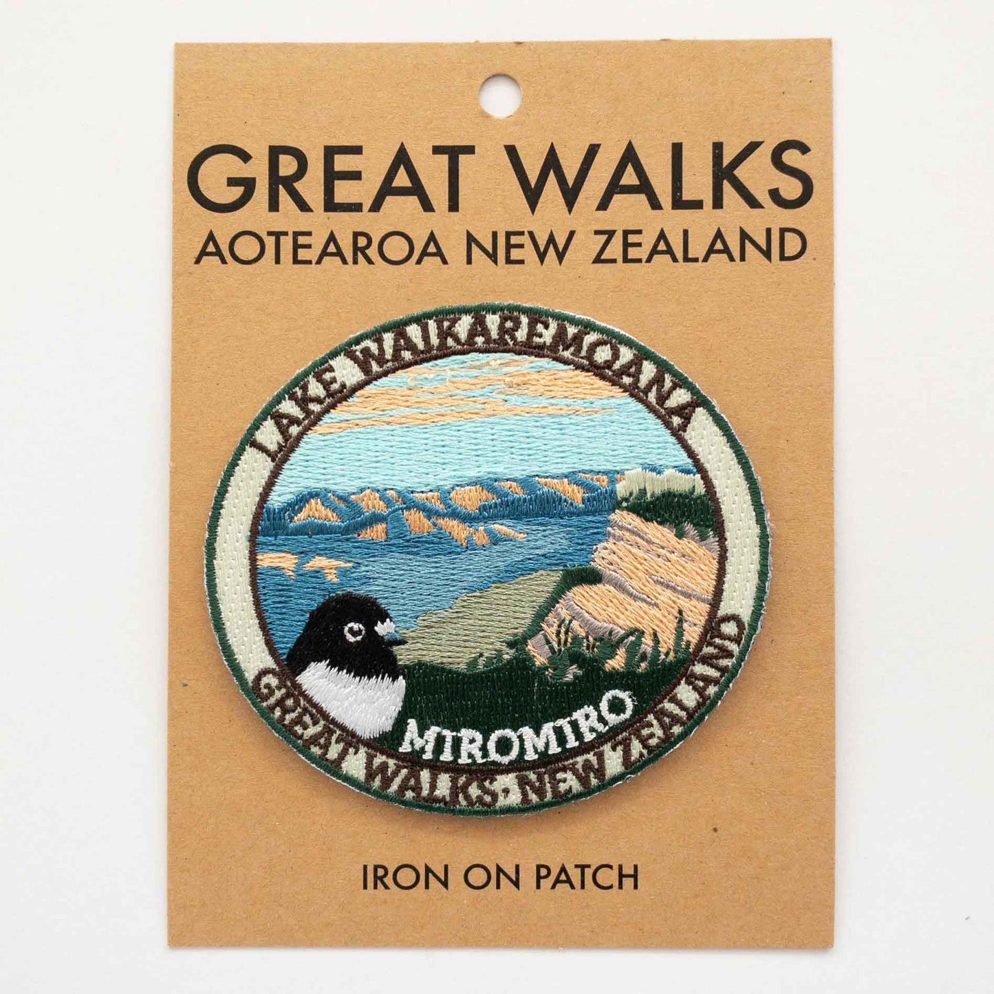 Oval, Embroidered Lake Waikaremoana Track patch, with a miromiro/tomtit bird, blue sky, hills and blue lake, on brown kraft backing card.