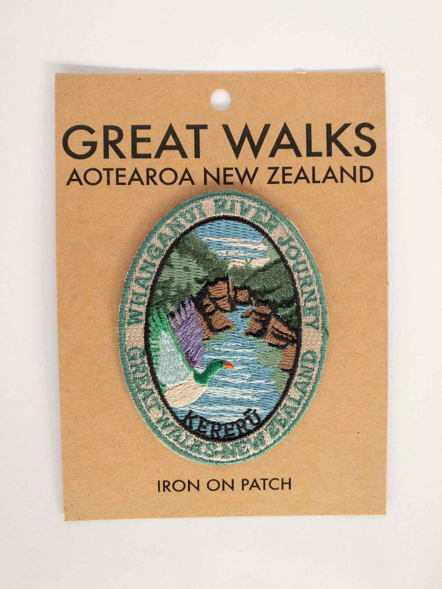 Oval, embroidered Whanganui River Journey Track patch, with a kereru/wood pigeon, green and brown banks and a blue river, on brown kraft backing card.