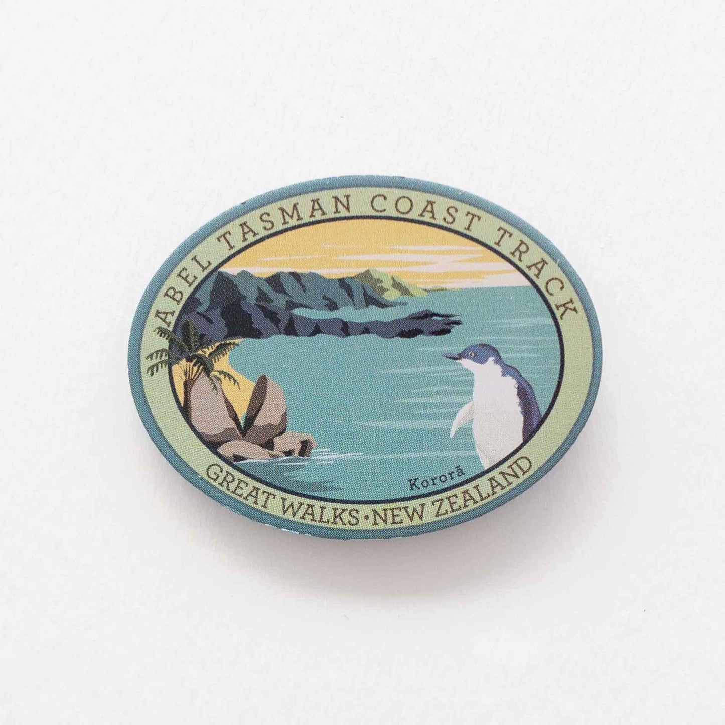 Oval Abel Tasman Coast Track patch, with a little penguin, split apple rock, yellow sky and sandy bays.