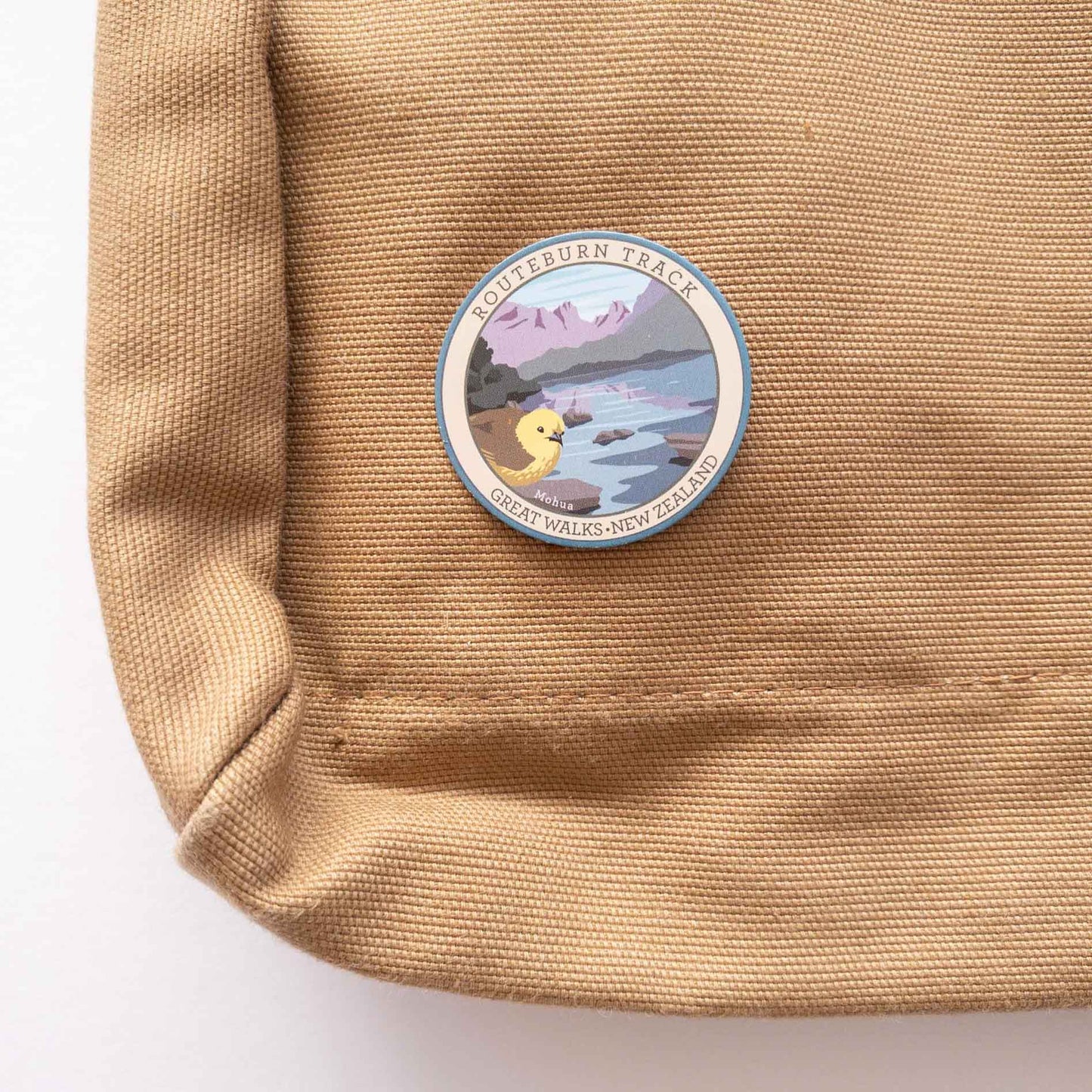 Oval Whanganui River Journey Track pin, with a kereru/wood pigeon, green and brown banks and a blue river, on a brown canvas bag.
