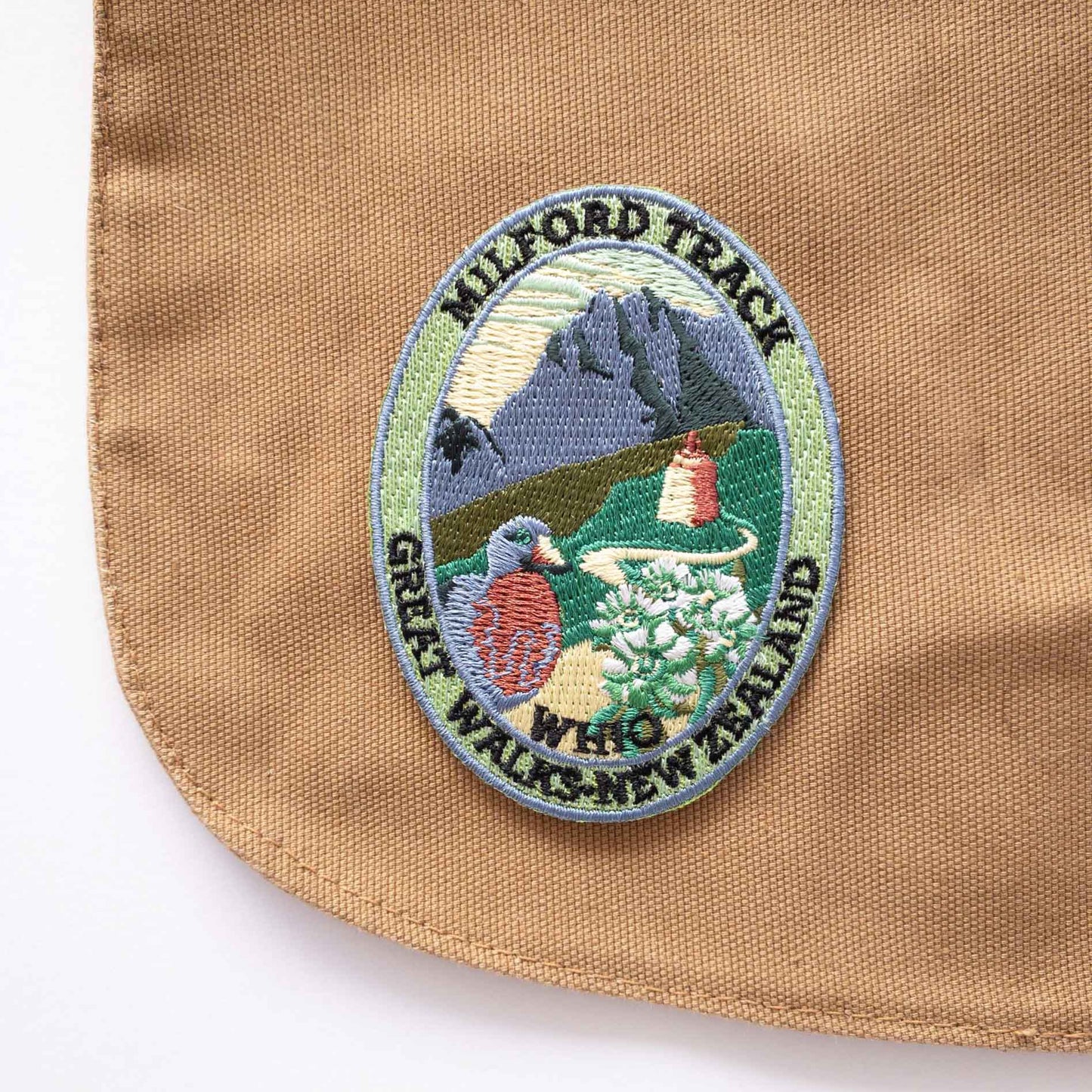 Oval, embroidered Milford Track patch, with a whio/blue duck, blue peak and mountain buttercup, on a brown canvas bag..