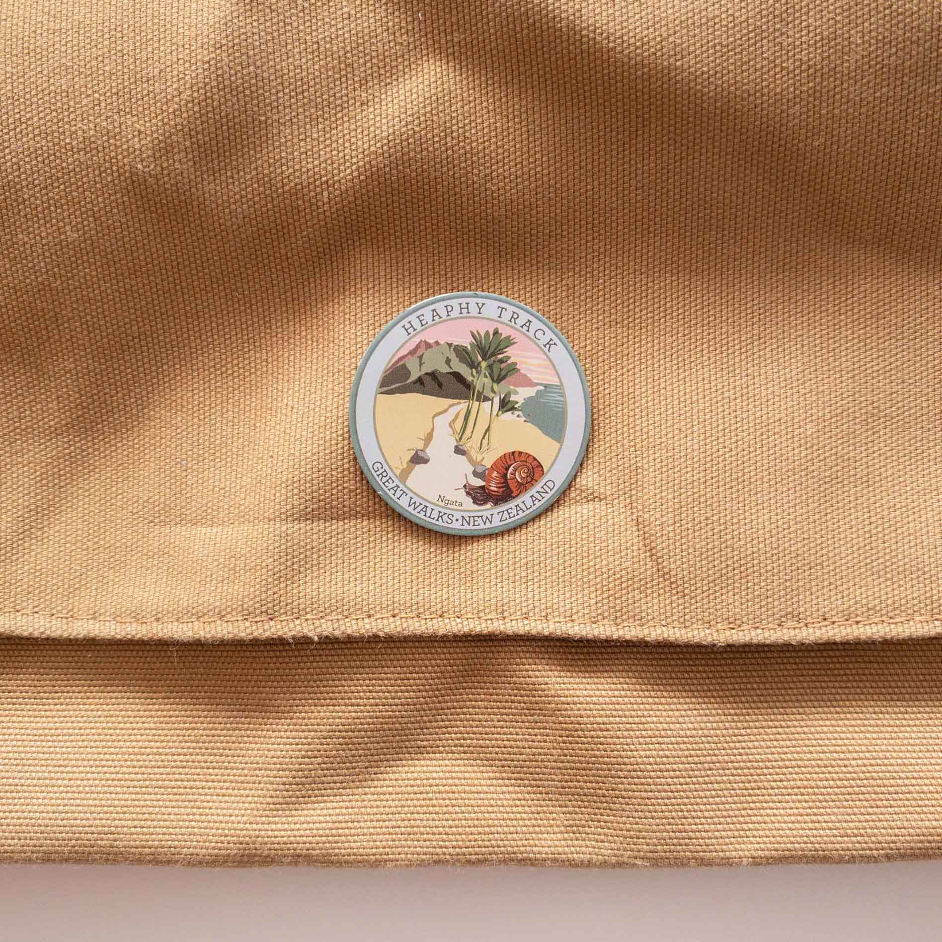 Round Heaphy Track pin, with a snail, nikau palms, green hills and pink sky, on a brown canvas bag.