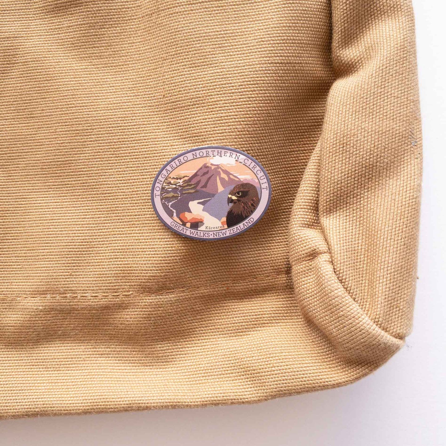 Oval Paparoa Track pin, with a weka, green hills, mountain daisy and tussock, on a brown canvas bag.